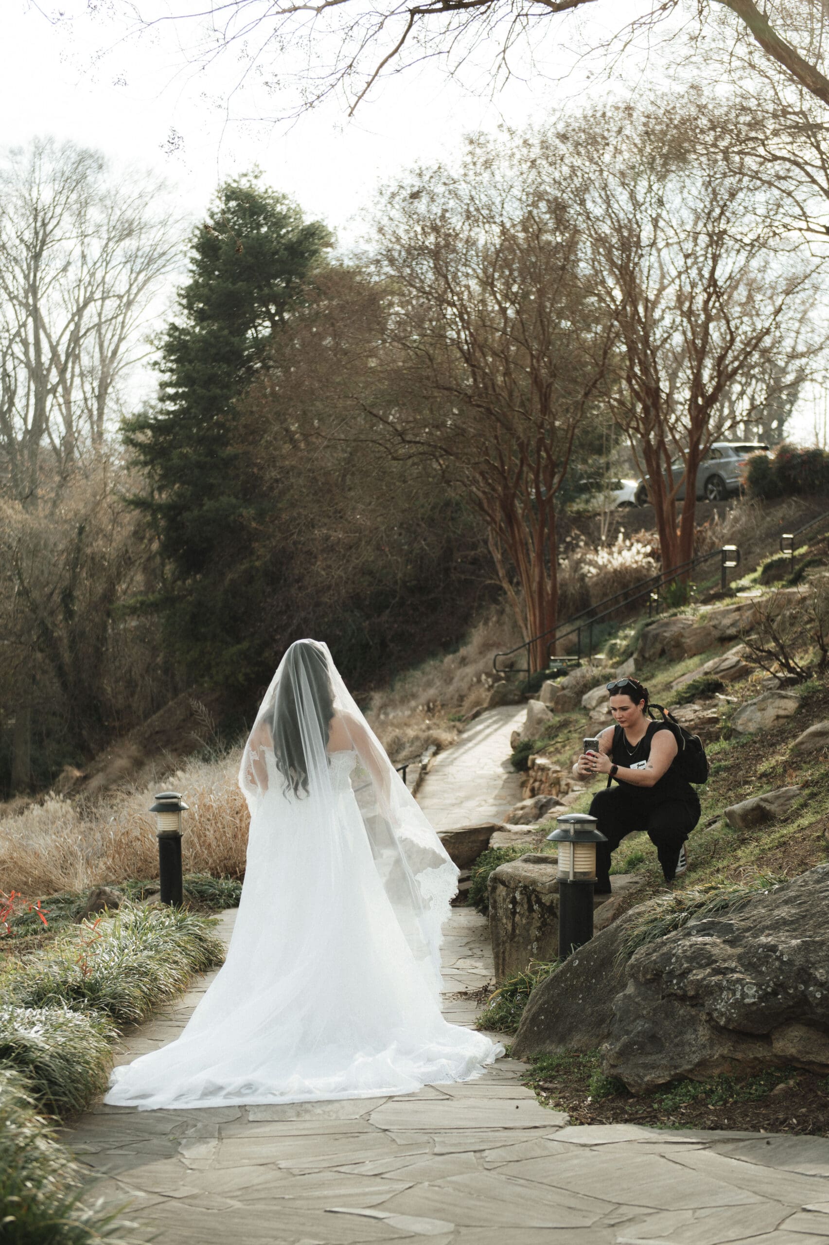 Wedding content creator BTS With Bonfante photographing a bride in a flowing white gown and veil along a stone pathway at Falls Park in downtown Greenville, capturing behind-the-scenes wedding content surrounded by trees and natural landscape.