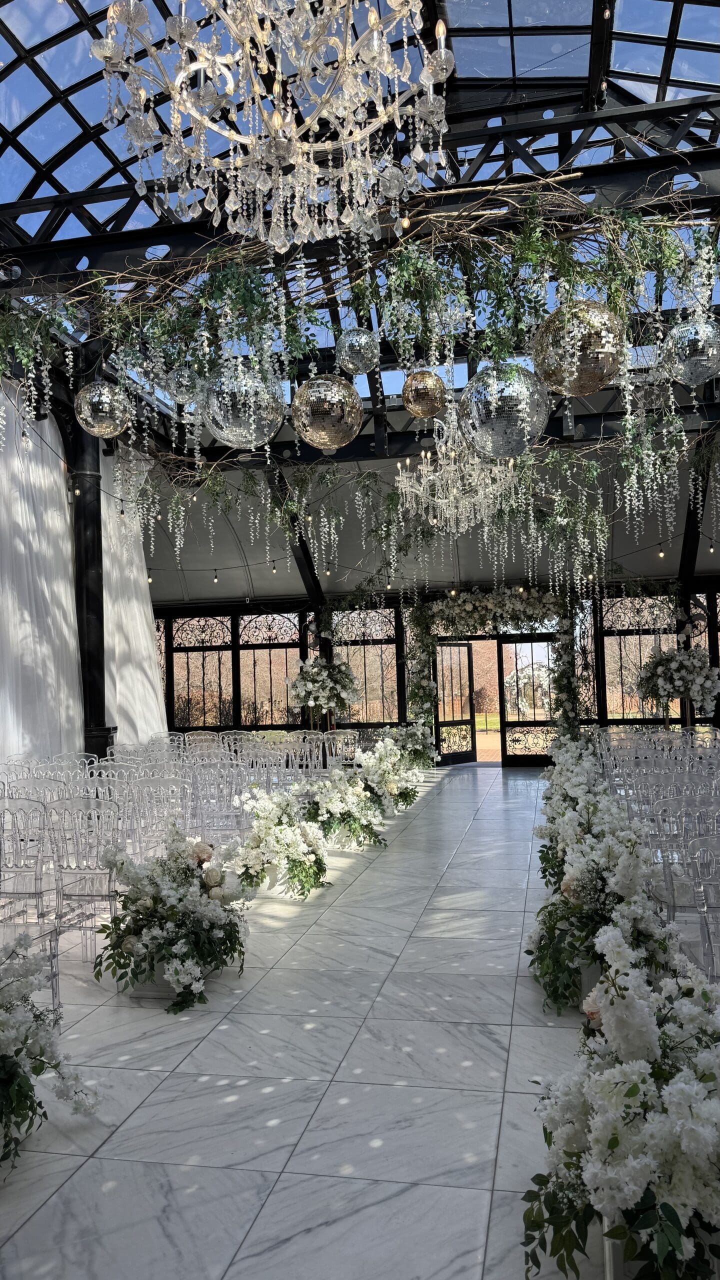 Wedding ceremony setup at Edinburgh West featuring a glass atrium with crystal chandeliers, hanging disco balls, and cascading greenery above a white marble aisle lined with lush white floral arrangements and clear ghost chairs, captured by wedding content creator BTS With Bonfante.
